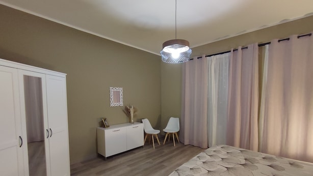 A bedroom with a bed, a wardrobe, a desk, two chairs, a hanging light fixture, and a window with curtains.