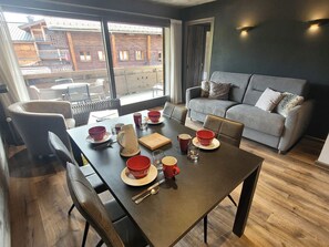 Ein Essbereich mit einem Tisch für vier Personen, einem grauen Sofa und Blick auf ein Holzhaus draußen.