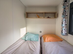 Une petite pièce avec deux lits, une étagère au-dessus et une fenêtre ornée de rideaux à motifs.