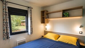 A bedroom with a bed, a window with curtains, a shelf with a box, and wall-mounted lights. - Mobile home 32m² with terrace, 3 bedrooms for 6 people (Granges-Aumontzey-Aumontzey)