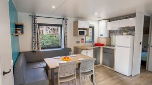 A compact living space with a kitchenette, a dining area with a table and chairs, a sofa, and a window with curtains. - Mobile home 32m² with terrace, 3 bedrooms for 6 people (Granges-Aumontzey-Aumontzey)