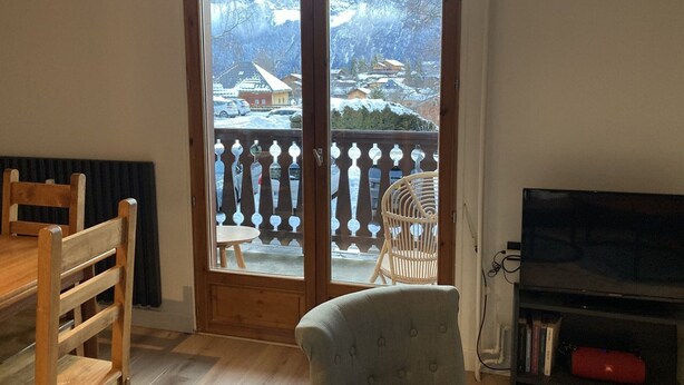 Ein Zimmer mit Esstisch aus Holz, einem gepolsterten Sessel, einem Fernseher und einem Balkon mit Blick auf eine verschneite Landschaft und Häuser.