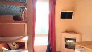 A room with bunk beds, a fireplace, a small table, and a window with curtains. - Modern and cozy studio at the foot of the slopes in Isola 2000 (Isola 2000)