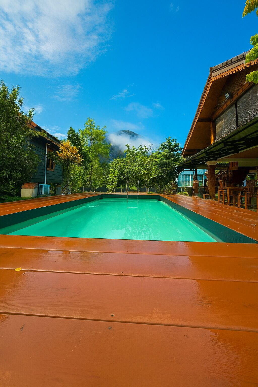 Una piscina con solárium de madera, rodeada de árboles y edificios de arquitectura tradicional.