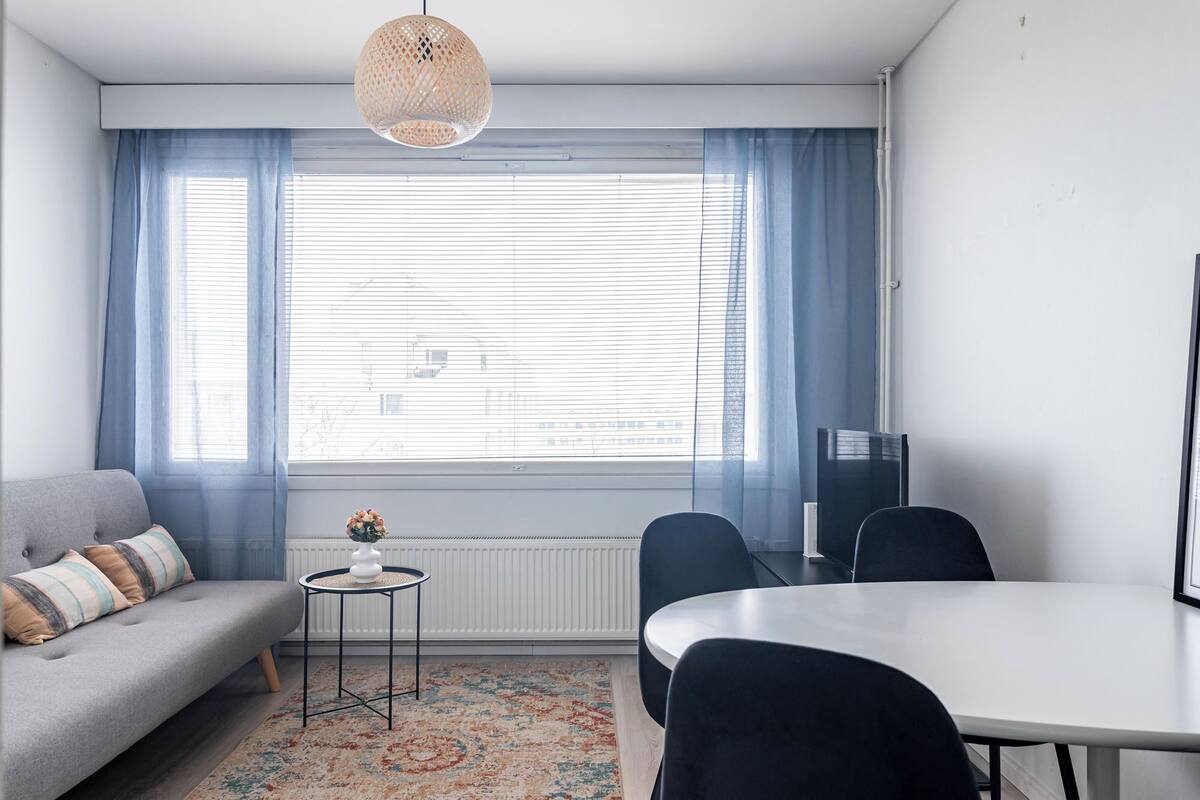 Een moderne woonkamer met een grijze bank, een klein rond tafeltje met een vaas, een eettafel met stoelen, en een groot raam met zonwering.