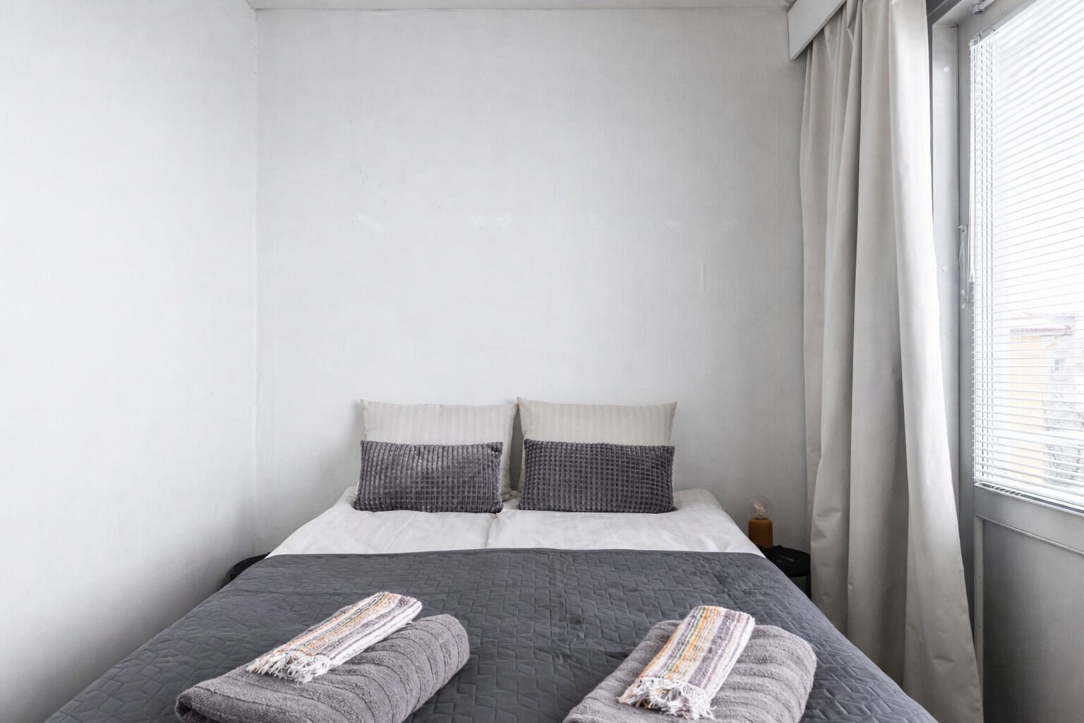 Un lit simple avec une couette grise et deux oreillers, une table de chevet avec une lampe, et une fenêtre avec des rideaux.
