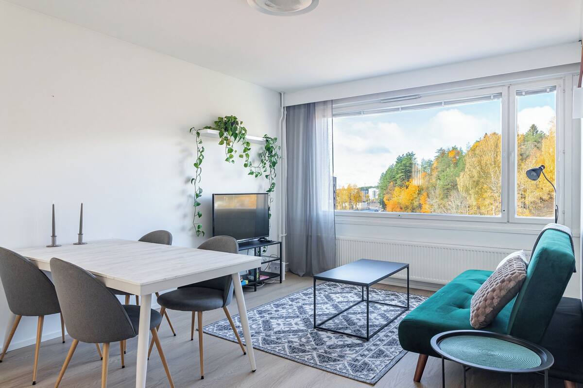 Een moderne woonkamer met een eethoek, een televisie en uitzicht op herfstbomen.