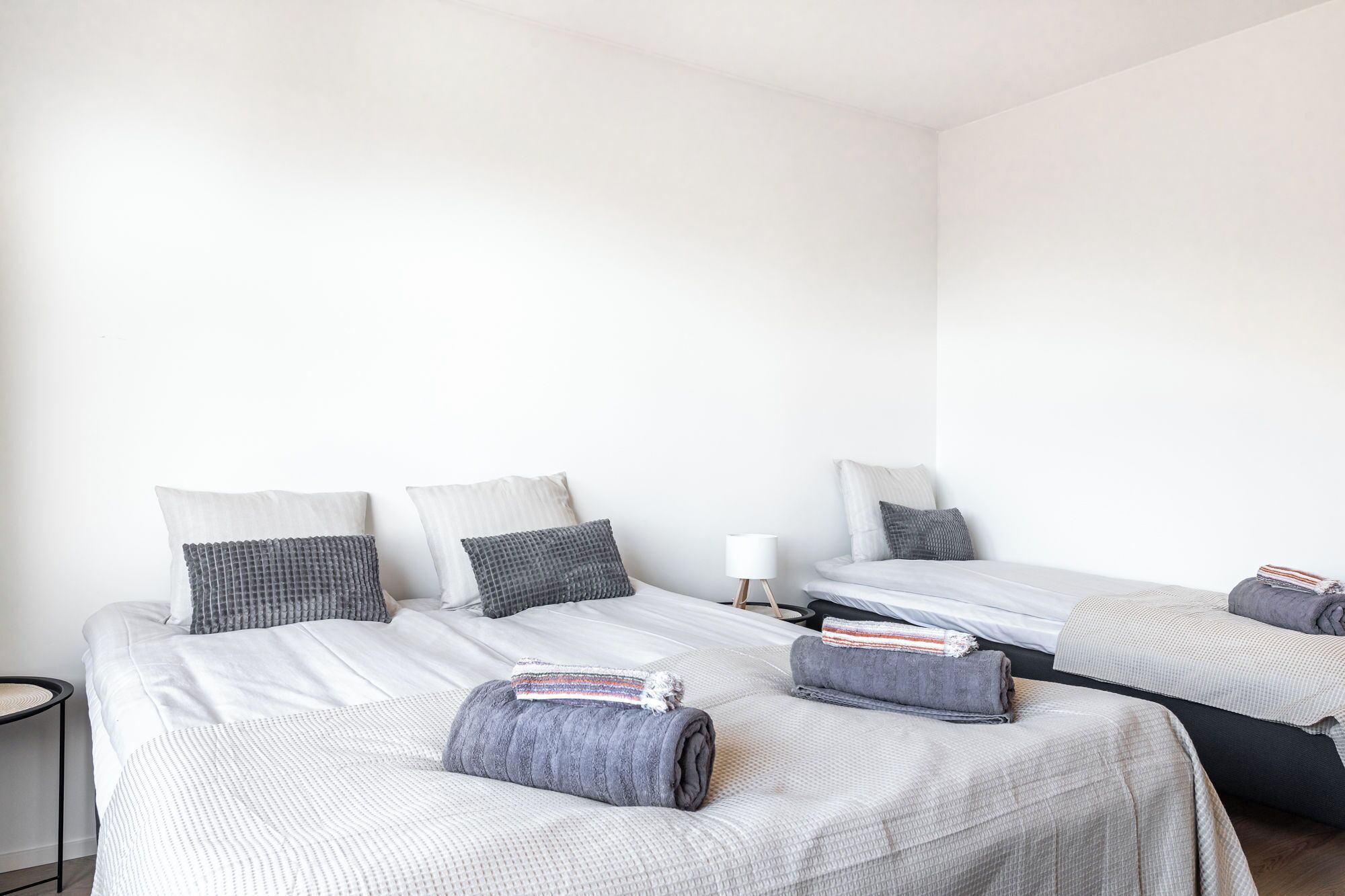 A hotel room with two beds, each with a grey blanket and pillows, a small bedside table with a lamp, and a chair.