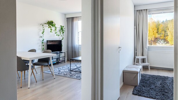 A modern living room with a flat-screen TV, a dining table with chairs, a small white stool, and a grey rug. - 1BR Apartments next to Railway Station, Free Parking, Balcony (Lahti)