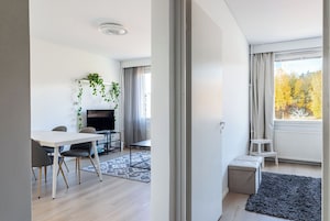 Una sala de estar moderna con un televisor de pantalla plana, una mesa de comedor con sillas, un taburete blanco pequeño y una alfombra gris.