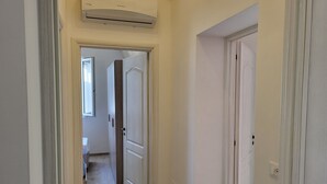 Un couloir étroit, avec une porte au fond, une fenêtre sur la gauche et un appareil de climatisation au-dessus.