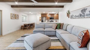 Een moderne woonkamer met een grijze modulaire bank, een poef en een vloerkleed met een patroon. Er is een eethoek met een tafel en stoelen, en een keuken op de achtergrond.