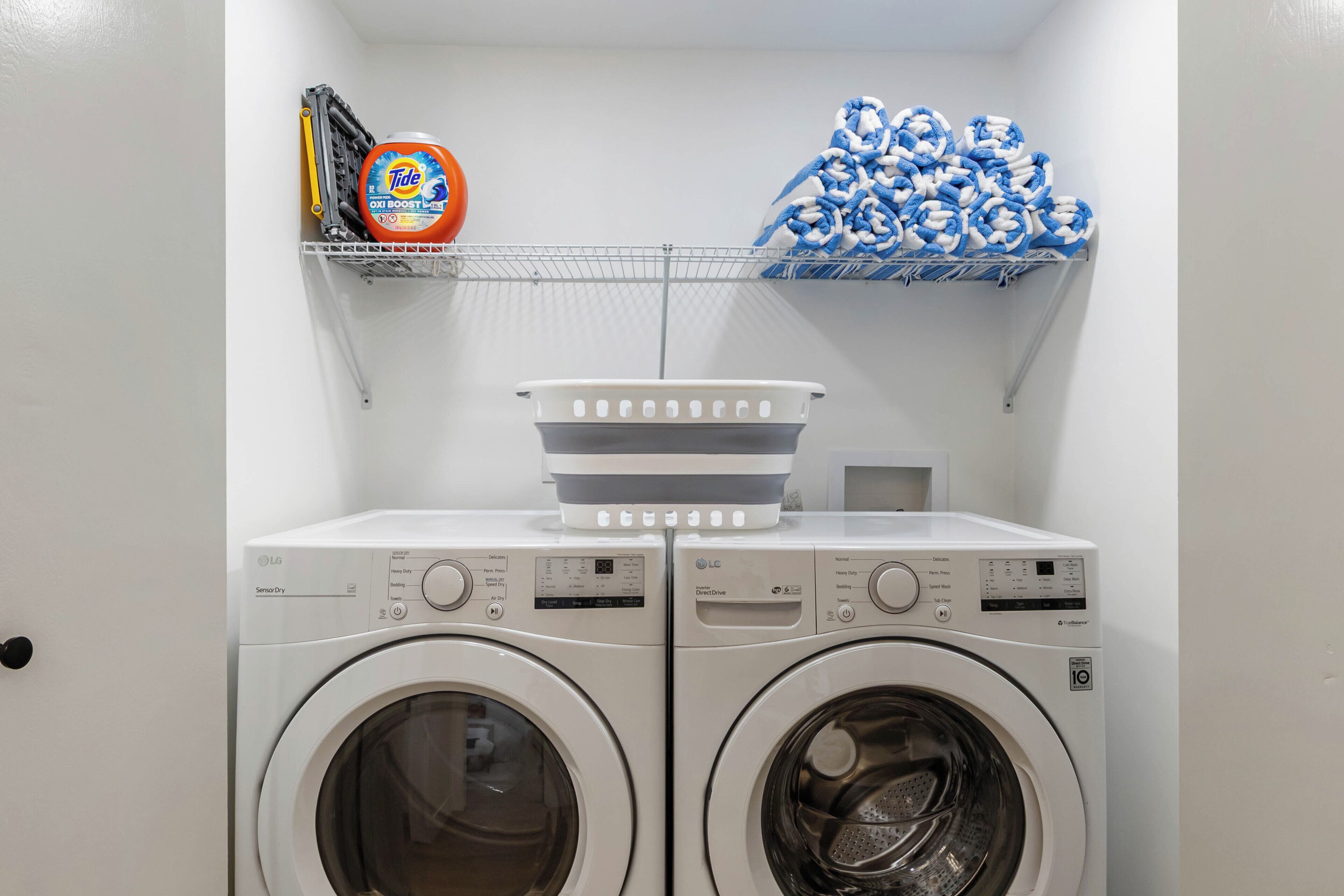 A laundry area with two front-loading washing machines, a detergent bottle, and a stack of towels on a shelf.