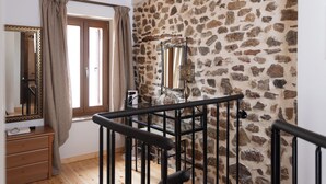 A room with a stone wall, a wooden ceiling, a mirror, a window with curtains, and a staircase with a metal railing. - To spiti sto castro (Nafpaktos)