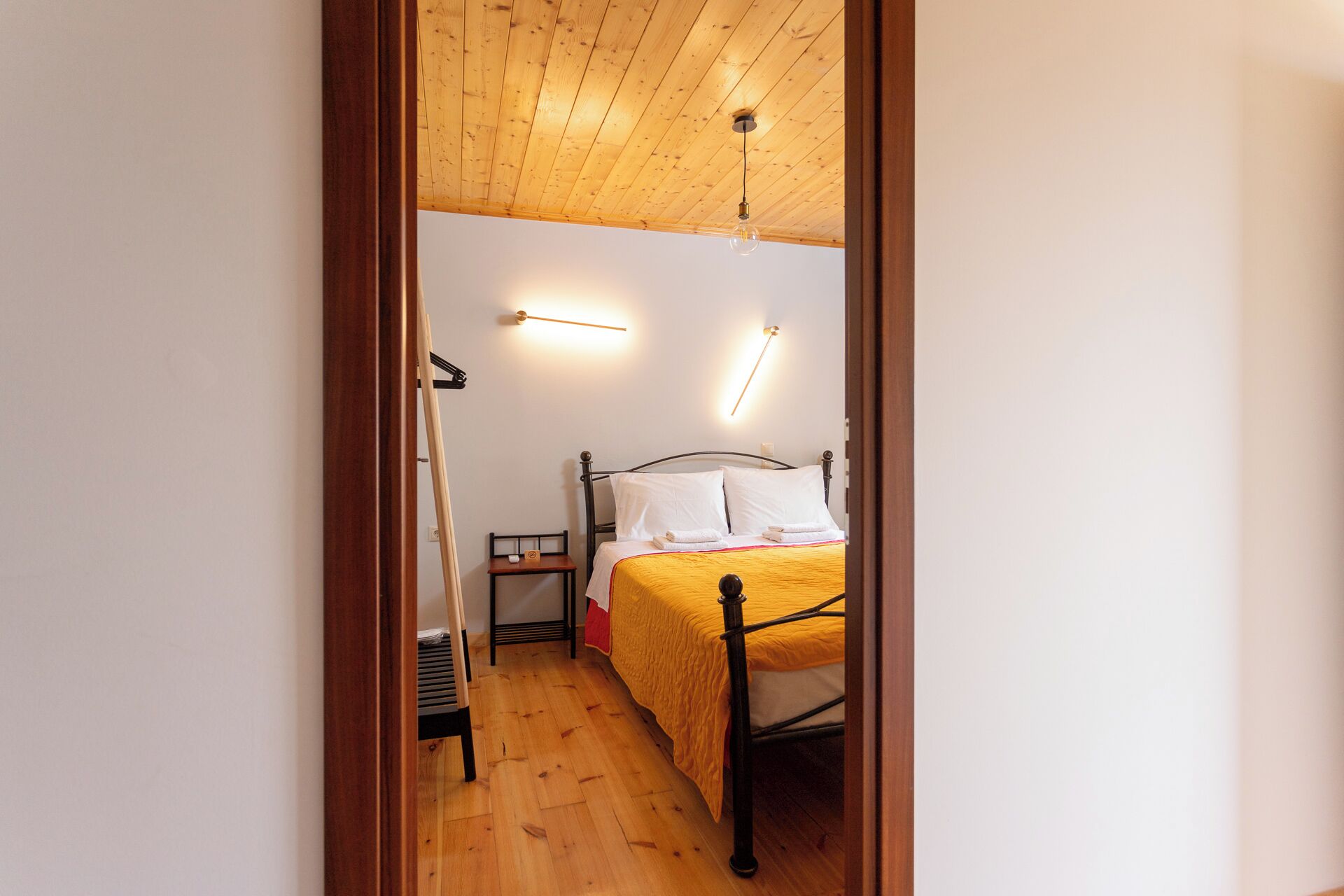 Een smalle gang die leidt naar een slaapkamer met een houten plafond, een bed met een gele sprei en een klein nachtkastje met een lamp.