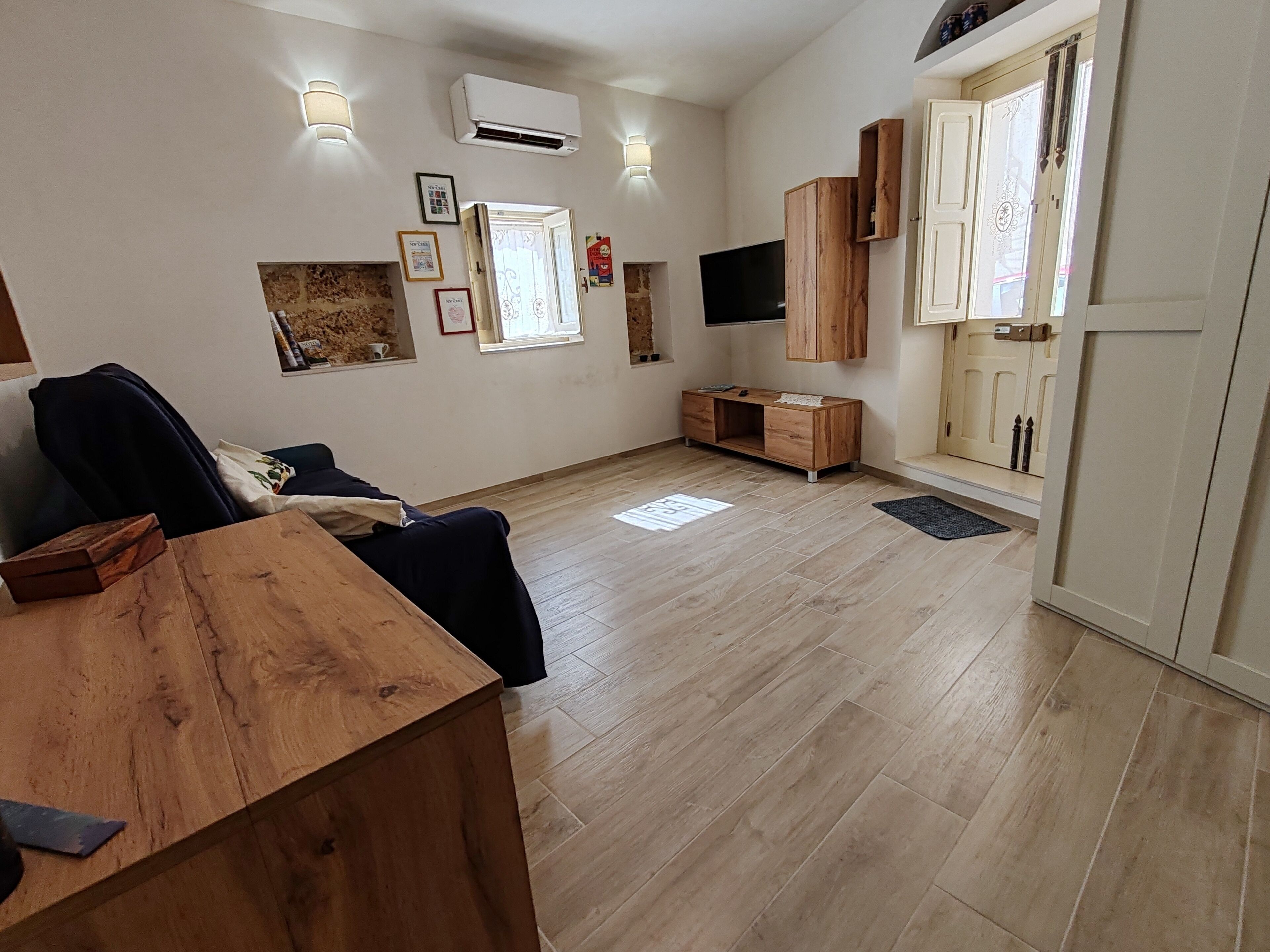 A spacious living area with a wooden table, a sofa, and a TV mounted on the wall. There are wall-mounted lights, a door with decorative glass panels, and a small cabinet.