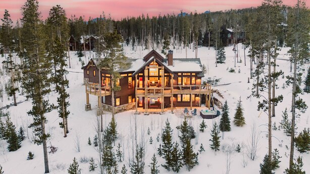 A large, multi-story lodge with a prominent central deck and multiple rooms, surrounded by a snowy forest.