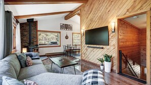 A modern living room with a large sofa, a coffee table, and a fireplace. There is a dining area with a table and chairs, and a TV mounted on the wall.