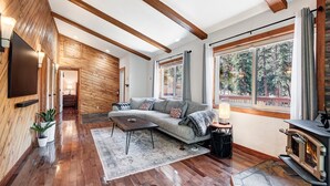 A modern living room with a wooden floor, a grey sofa, a fireplace, and a large window with a view of trees.