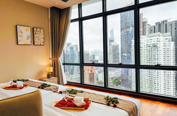 Una habitación de hotel con un ventanal grande, una cama con cubrecama blanco, una mesita de noche con lámpara y una bandeja con pétalos de rosa y una copa de vino.
