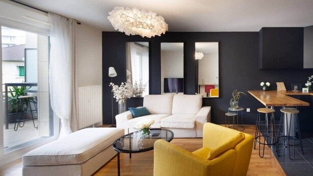 Un salon moderne avec un canapé, un fauteuil jaune, une table basse en verre et un coin repas comprenant une table en bois et des tabourets de bar.