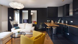 Un salon moderne avec un canapé, un fauteuil jaune et une table basse en verre. On aperçoit également un coin cuisine doté d’un plan de travail en bois et de tabourets.
