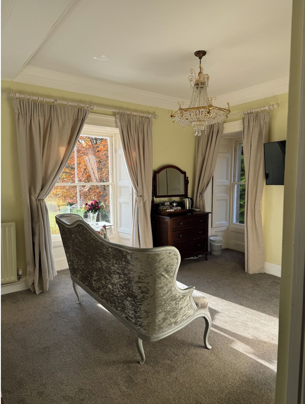 A room with a large window, a chandelier, a vintage armchair, and a dresser.