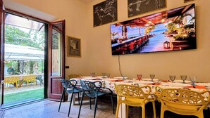 A dining area with a long table, chairs, and a television screen displaying a beach scene. - Leonardo House Pisa Garden Center Whirlpool Pool (Pisa)