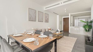 A modern dining area with a wooden table set for four, grey upholstered chairs, and a kitchen in the background.