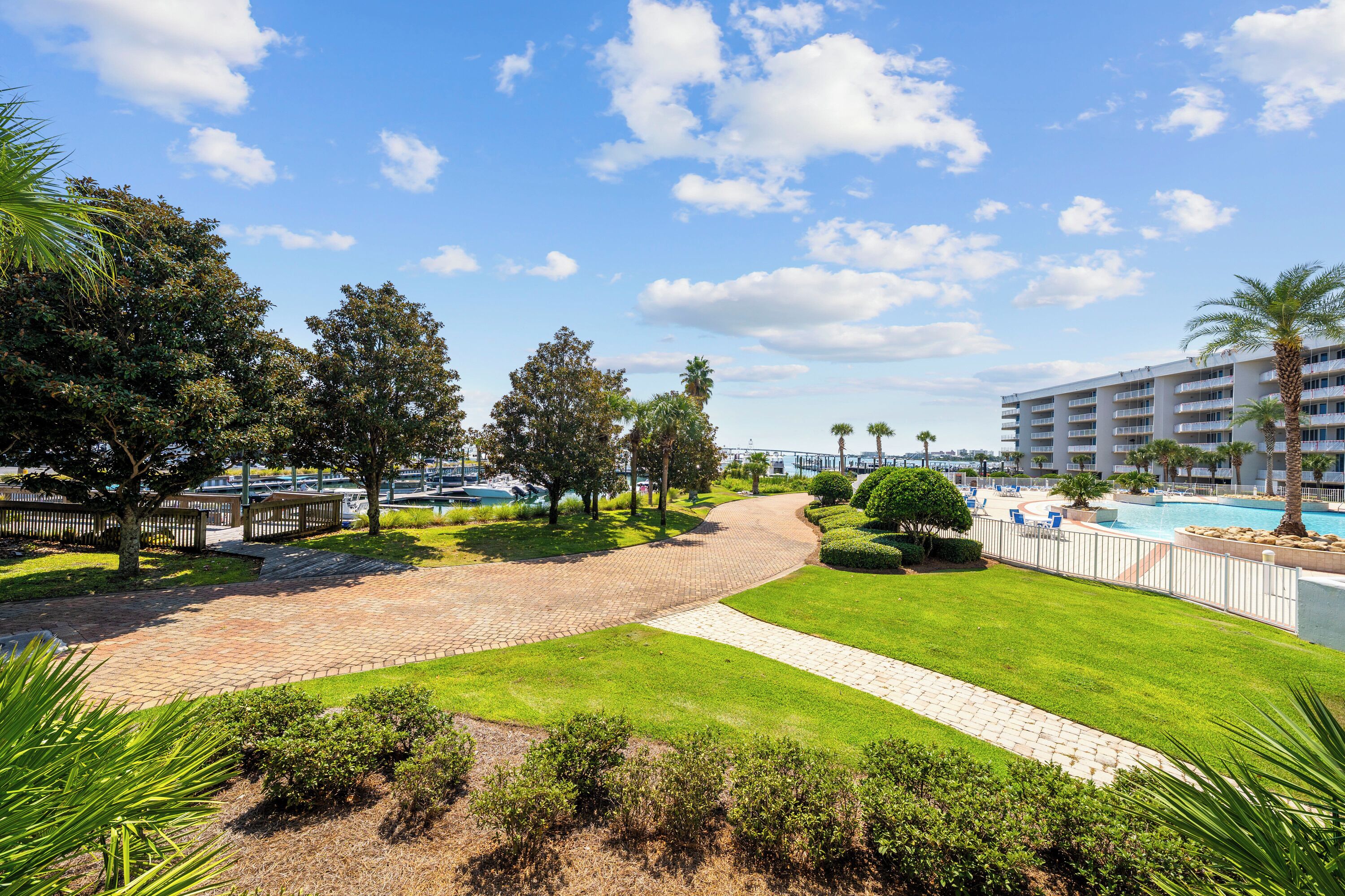 Un sendero pavimentado que conduce a una zona de piscina con palmeras y un hotel de varios pisos al fondo.