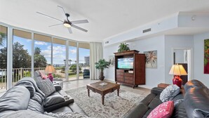 A spacious living room with a large sofa, a coffee table, and a television. There is a balcony with a view of palm trees and buildings. - The Moorings 101 (Orange Beach)