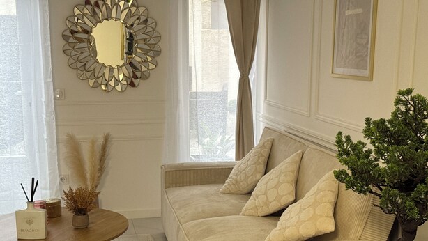 Ein Wohnzimmer mit einem beigefarbenen Sofa, einem runden Tisch mit einer Schale Orangen, einer Topfpflanze und einem dekorativen Spiegel an der Wand.