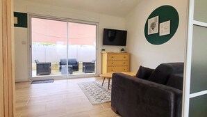 A living room with a sofa, a TV, a wooden cabinet, and a sliding glass door leading to an outdoor area with chairs and a table. - STUDIO 30 M2 (Labastide-Saint-Sernin)