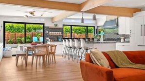 A modern kitchen with white cabinets, a dining area with wooden chairs, and large windows with a view of greenery. - The Spacious Open Palms (Palm Springs)