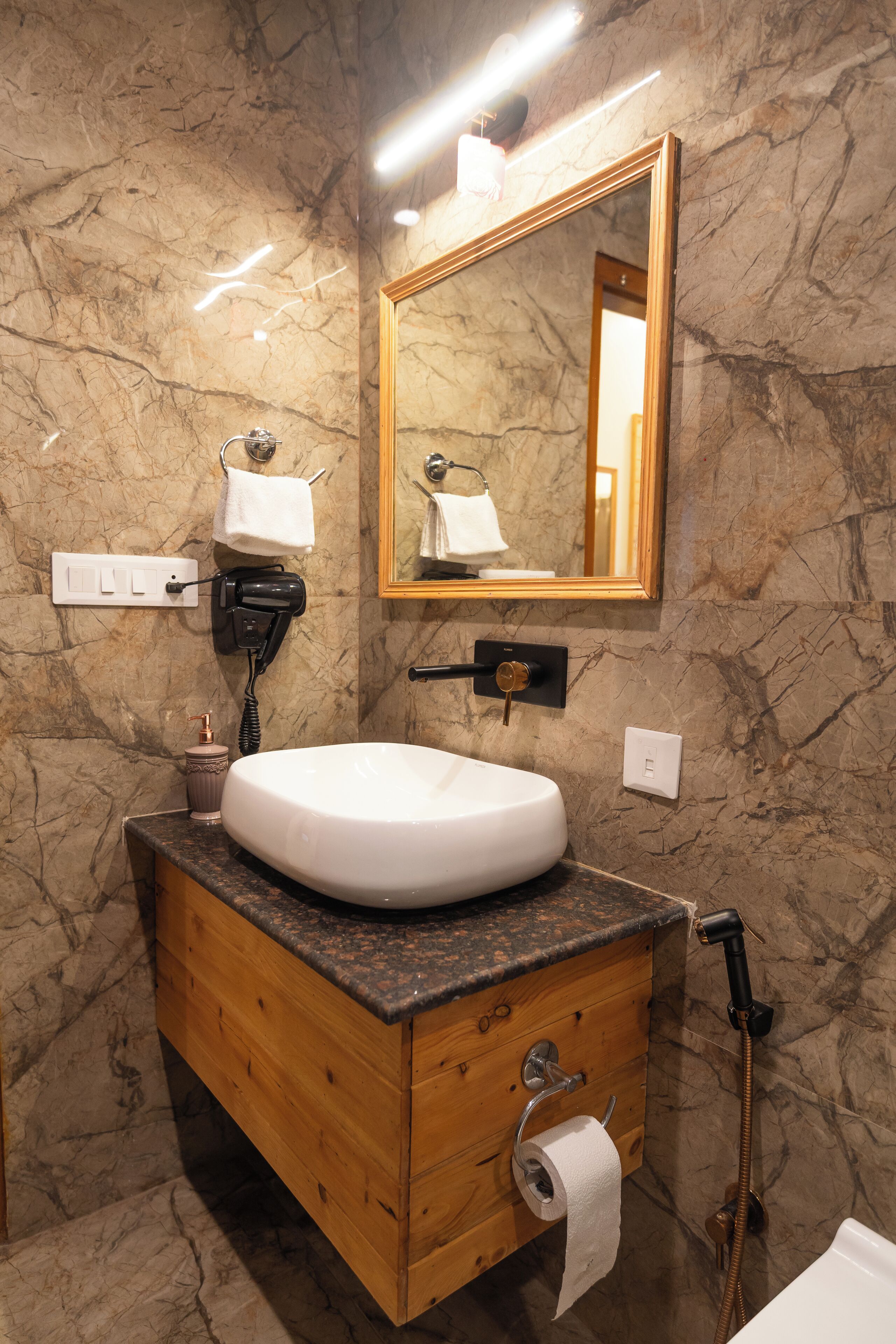 Un bagno con una parete in marmo, un mobile lavabo in legno, un lavandino bianco, un porta asciugamani, uno specchio e un dispenser per asciugamani.
