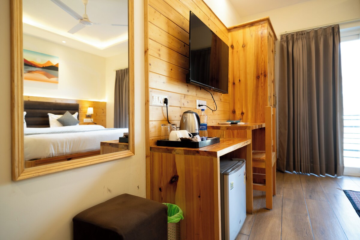 A compact hotel room with a bed, a desk with a television, a small kitchenette, and a large mirror.