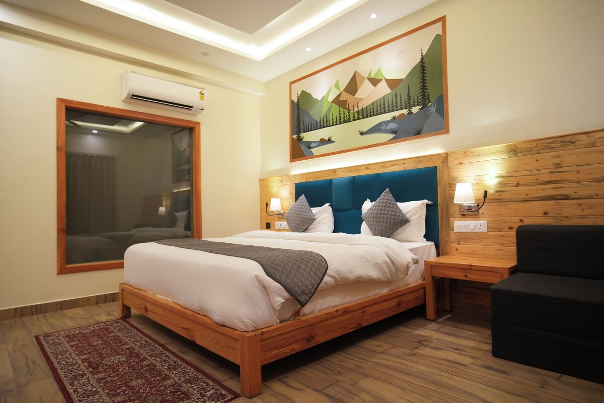 A hotel room with a wooden bed, a large window, an air conditioning unit, a painting of mountains, and a wooden headboard.