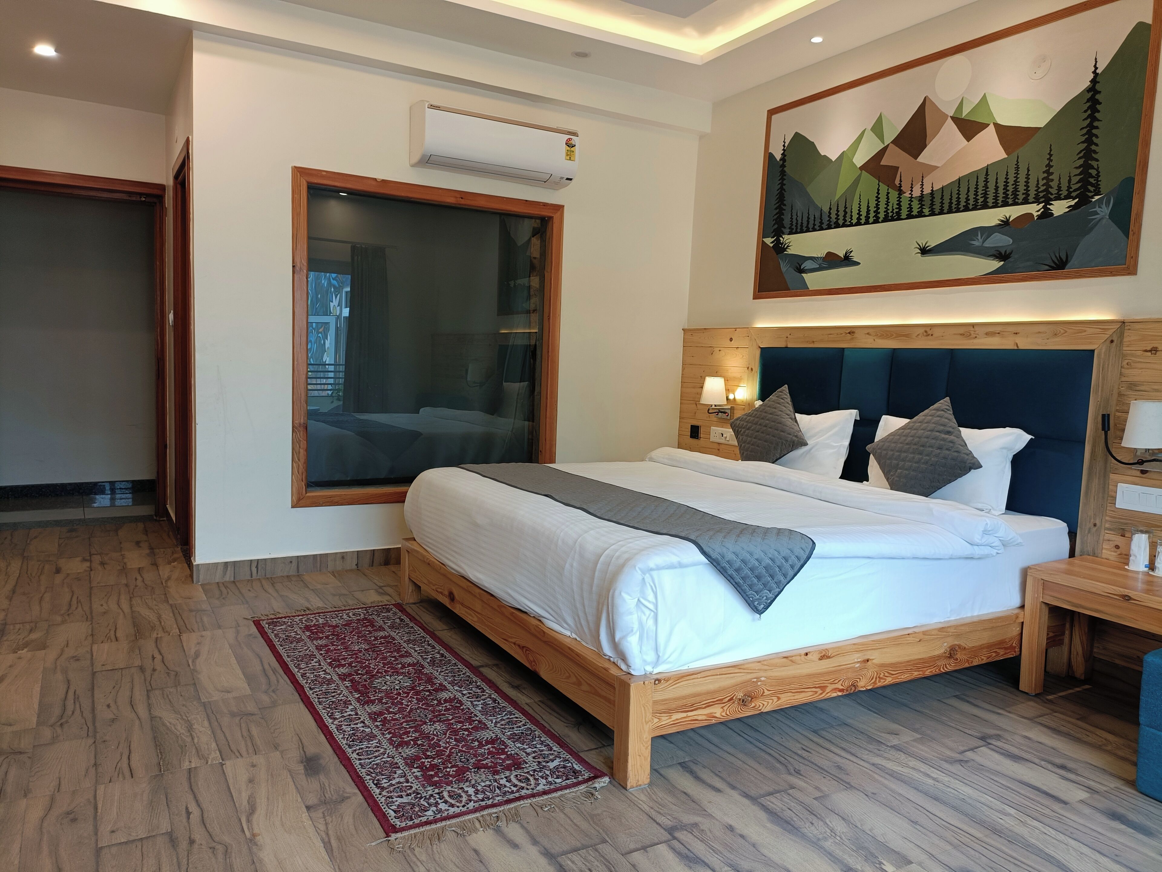 A hotel room with a large bed, wooden headboard, a window with a view, a painting on the wall, and a red rug.