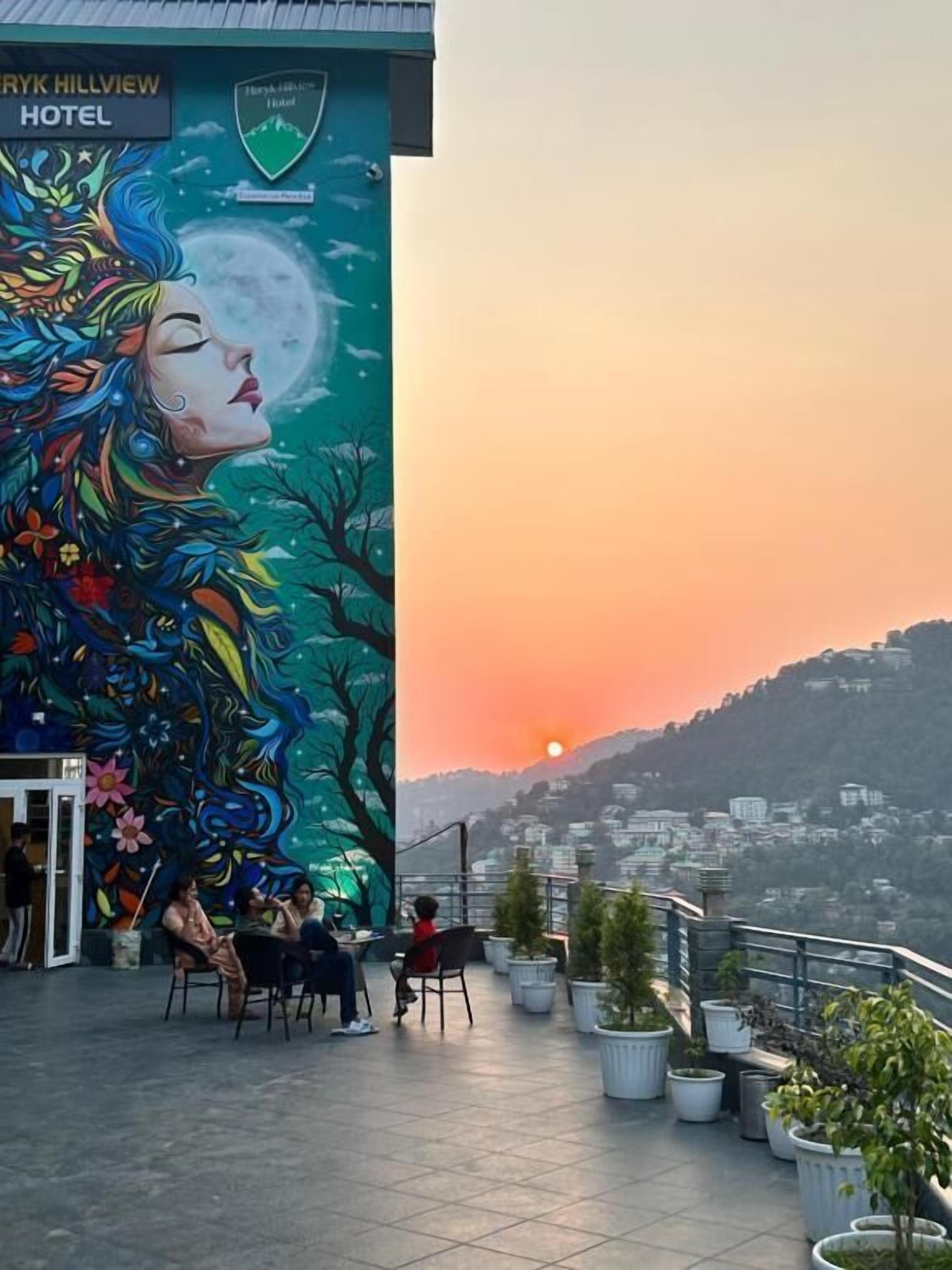 Exterior de un hotel con un mural de una mujer y árboles, mobiliario exterior y plantas en macetas.