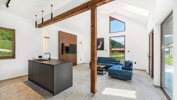 Ein modernes Interieur mit Kücheninsel, blauem Sofa und großem Fenster mit Blick ins Grüne.