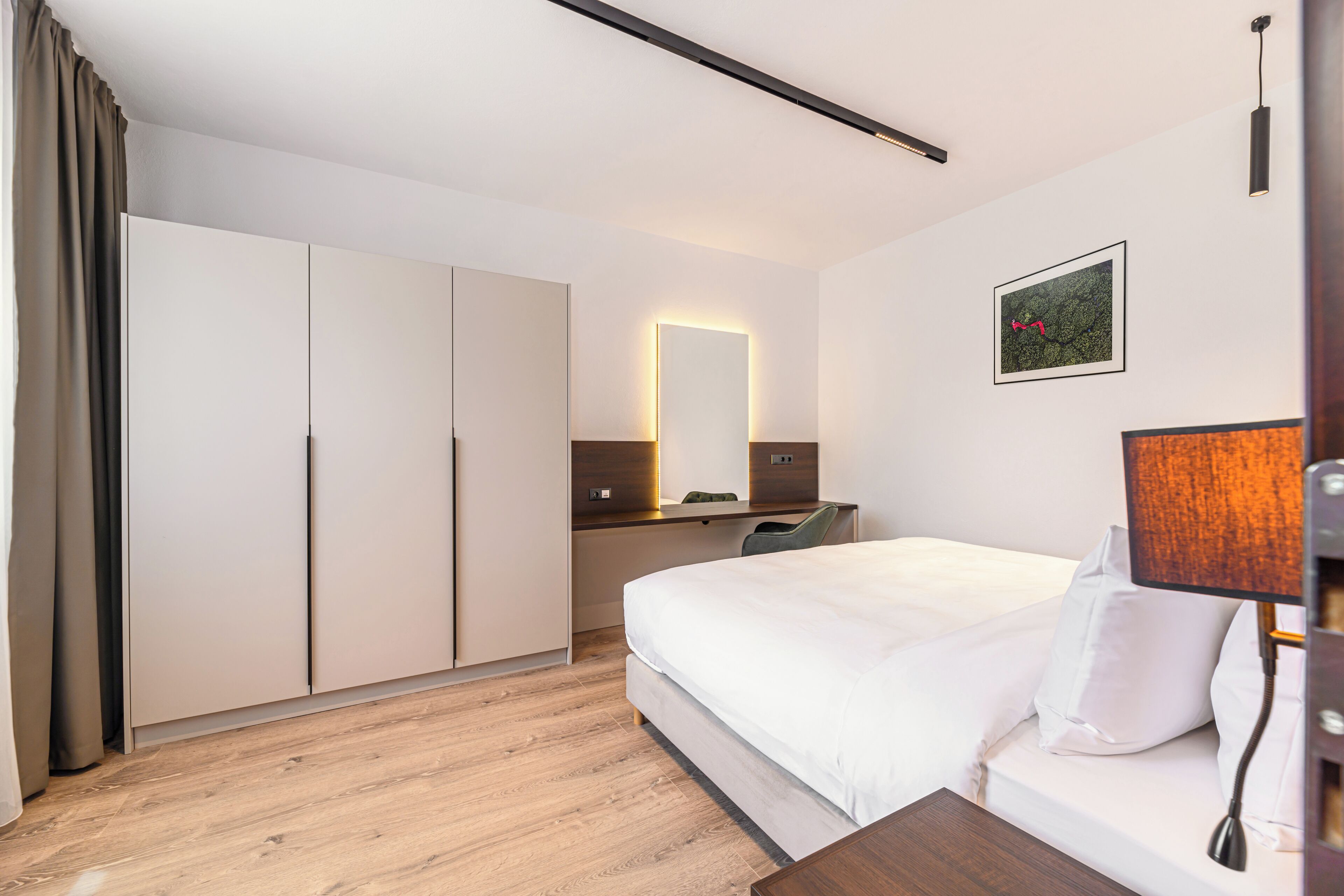 A modern hotel room with a large bed, a desk with a chair, a wardrobe, and a wall-mounted lamp.