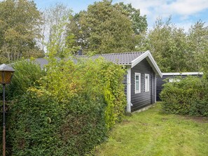 A small, dark-colored house with a sloped roof and white trim, surrounded by dense greenery and a well-maintained lawn.