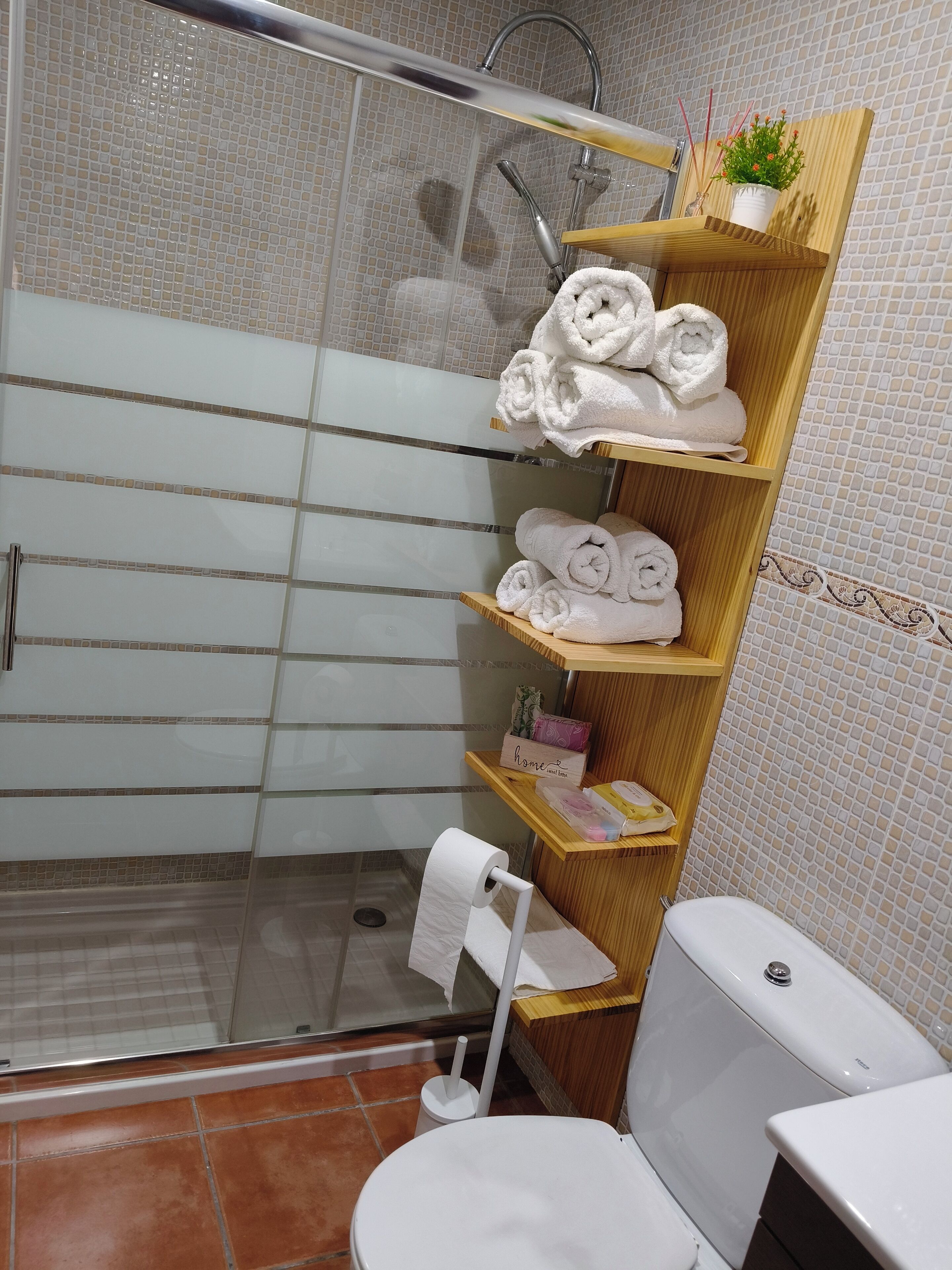 A bathroom with a glass shower enclosure, a toilet, and a shelf with towels and toiletries.
