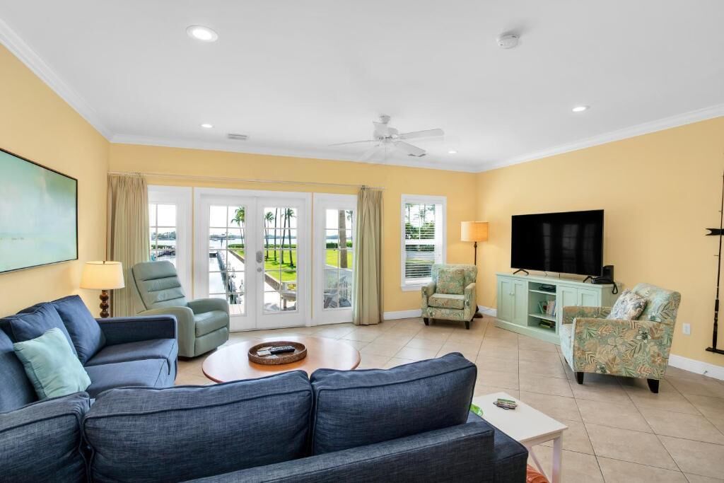 A living room with a sofa, armchairs, a TV, and a balcony with a view of palm trees.