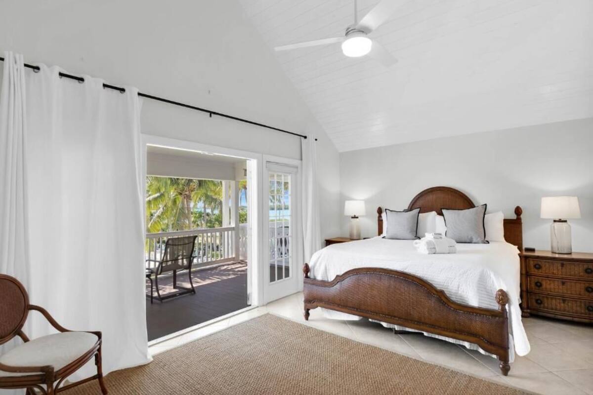 A bedroom with a large bed, a wooden nightstand, a chair, and a balcony with a view of palm trees.
