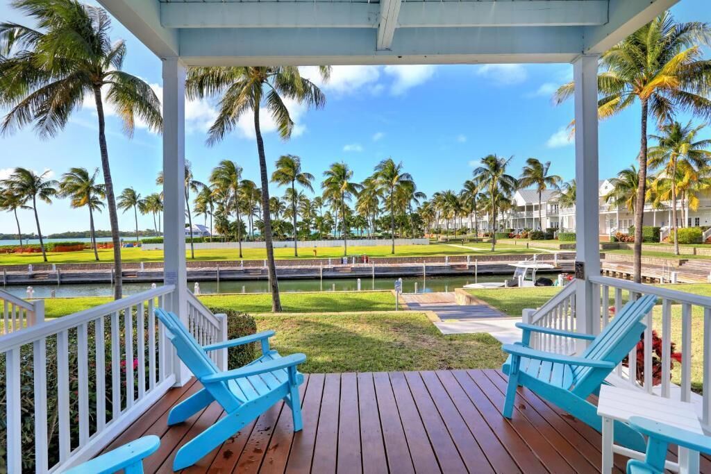 A covered deck with two blue Adirondack chairs, a view of a canal, and palm trees.