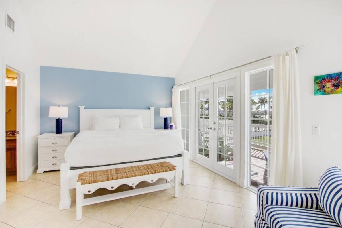 A bedroom with a bed, bedside tables, a bench, a striped sofa, and a balcony with glass doors.