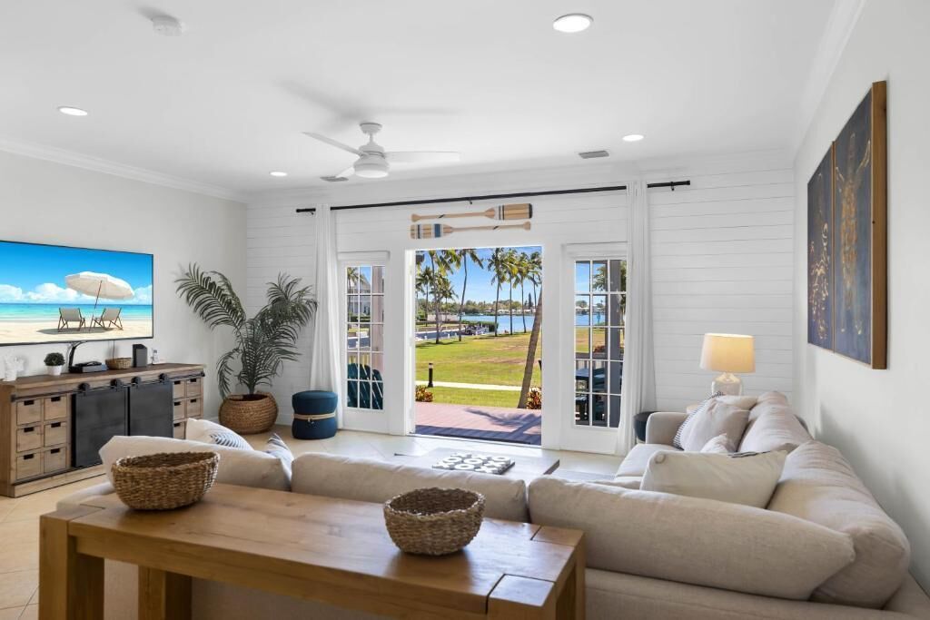 A modern living room with a television, a wooden table, and a sofa. There is a view of a pool and palm trees through the glass doors.