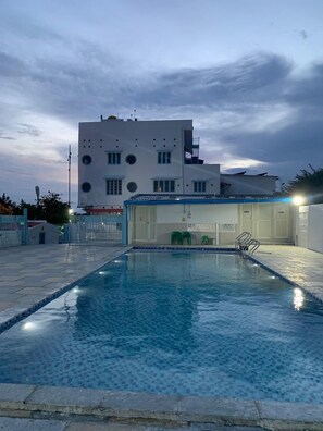 En swimmingpool med ett kaklat poolområde, en överbyggd del med stolar och en byggnad med modern design i bakgrunden.