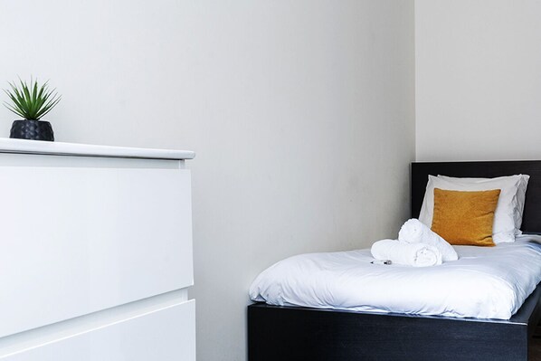 A modern hotel room with a bed, white bedding, a black headboard, a mustard-colored pillow, and a small potted plant on a white dresser.
